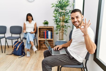 Téléchargez les photos : Les jeunes assis dans la salle d'attente travaillant avec un ordinateur portable faisant signe ok avec les doigts, sourire geste amical excellent symbole - en image libre de droit