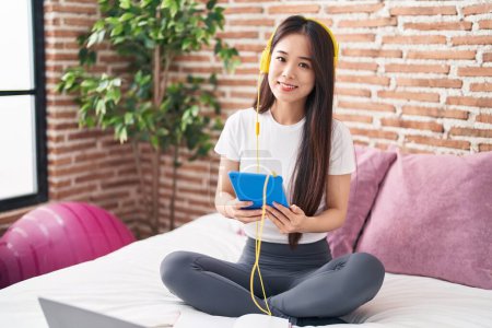 Téléchargez les photos : Jeune femme chinoise utilisant touchpad et écouteurs assis sur le lit dans la chambre - en image libre de droit