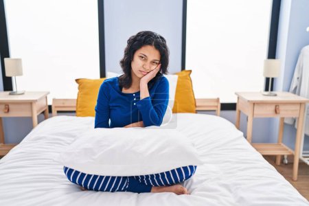 Foto de Mujer hispana con cabello oscuro sentada en la cama en casa pensando que se ve cansada y aburrida con problemas de depresión con los brazos cruzados. - Imagen libre de derechos