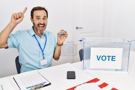 Téléchargez les photos : Homme d'âge moyen avec la barbe assis par bulletin de vote tenant i insigne vote sourire étonné et surpris et pointant vers le haut avec les doigts et les bras levés. - en image libre de droit