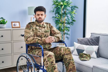 Photo for Arab man wearing camouflage army uniform sitting on wheelchair puffing cheeks with funny face. mouth inflated with air, crazy expression. - Royalty Free Image