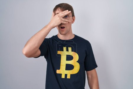 Photo for Caucasian blond man wearing bitcoin t shirt peeking in shock covering face and eyes with hand, looking through fingers with embarrassed expression. - Royalty Free Image