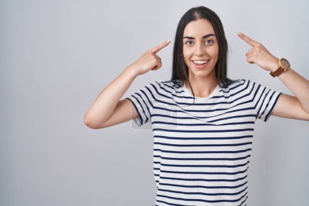Téléchargez les photos : Jeune femme brune portant un t-shirt rayé souriant pointant vers la tête avec les deux doigts des mains, bonne idée ou pensée, bonne mémoire - en image libre de droit