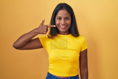 Téléchargez les photos : Jeune femme indienne debout sur fond jaune pointant avec le doigt de la main sur le visage et le nez, souriant gai. concept de beauté - en image libre de droit