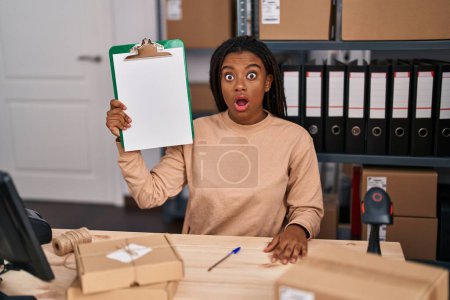 Photo for Young african american with braids working at small business ecommerce showing clipboard scared and amazed with open mouth for surprise, disbelief face - Royalty Free Image