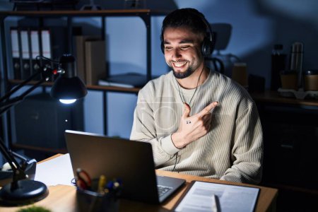 Téléchargez les photos : Jeune homme beau travaillant à l'aide d'un ordinateur portable la nuit joyeuse avec un sourire du visage pointant avec la main et le doigt sur le côté avec une expression heureuse et naturelle sur le visage - en image libre de droit