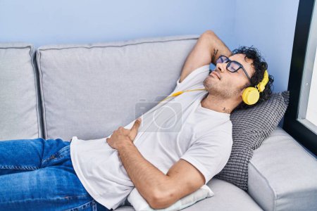 Téléchargez les photos : Jeune homme hispanique écoutant de la musique allongée sur le canapé à la maison - en image libre de droit