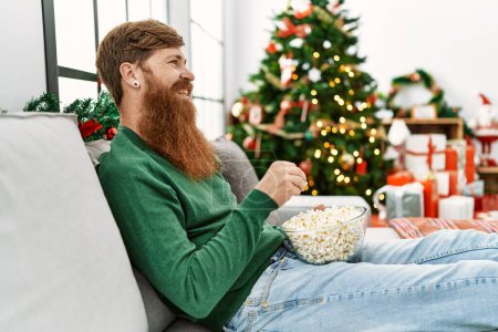 Photo for Young redhead man watching movie sitting by christmas tree at home - Royalty Free Image