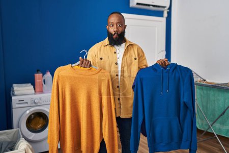 Photo for African american man holding clean clothes on hangers at laundry room making fish face with mouth and squinting eyes, crazy and comical. - Royalty Free Image