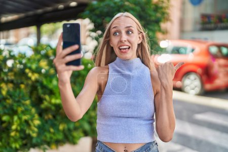 Téléchargez les photos : Young caucasian woman doing video call with smartphone pointing thumb up to the side smiling happy with open mouth - en image libre de droit