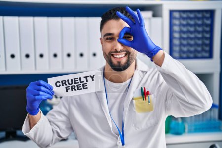 Foto de Joven hombre guapo que trabaja en el laboratorio científico sosteniendo crueldad bandera libre sonriendo feliz haciendo signo de ok con la mano en el ojo mirando a través de los dedos - Imagen libre de derechos