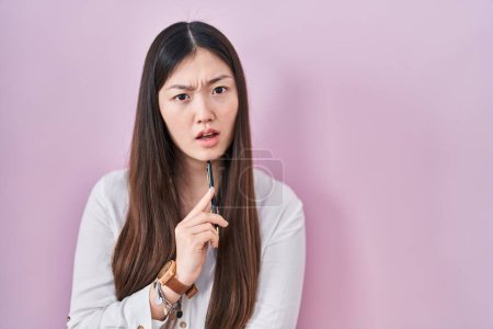 Foto de Mujer joven china sosteniendo lápiz sobre fondo rosa en la cara de choque, mirando escéptico y sarcástico, sorprendido con la boca abierta - Imagen libre de derechos
