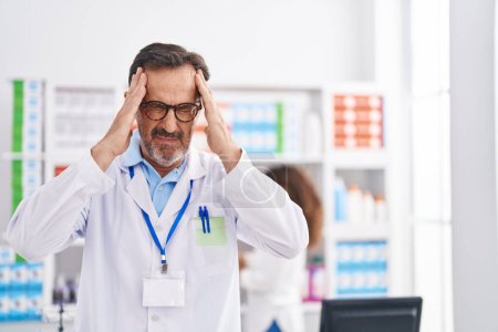 Foto de Hombre hispano de mediana edad que trabaja en farmacia con la mano en la cabeza, dolor de cabeza porque el estrés. padecer migraña. - Imagen libre de derechos