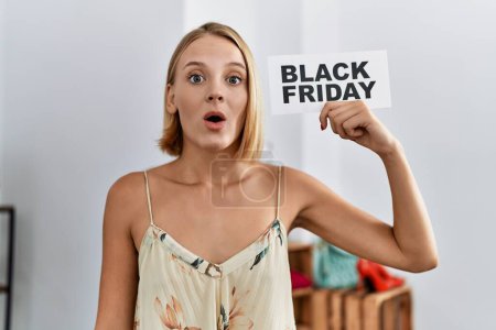 Téléchargez les photos : Jeune femme caucasienne tenant bannière vendredi noir au magasin de détail effrayé et étonné avec la bouche ouverte pour la surprise, visage incrédule - en image libre de droit