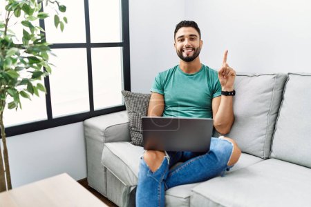 Photo for Young handsome man with beard using computer laptop sitting on the sofa at home showing and pointing up with finger number one while smiling confident and happy. - Royalty Free Image
