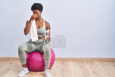 Foto de Mujer afroamericana usando ropa deportiva sentada en una pelota de pilates cansada frotando la nariz y los ojos sintiendo fatiga y dolor de cabeza. concepto de estrés y frustración. - Imagen libre de derechos
