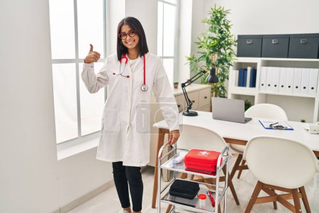 Foto de Mujer hispana joven vistiendo uniforme médico y estetoscopio sonriendo feliz y positivo, pulgar hacia arriba haciendo excelente y signo de aprobación - Imagen libre de derechos