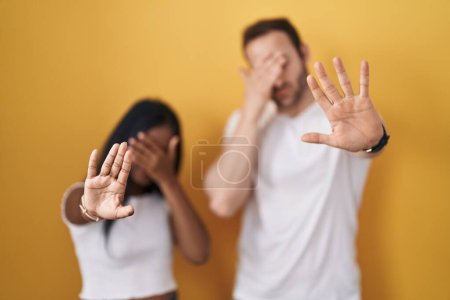 Ein gemischtrassiges Paar, das vor gelbem Hintergrund steht und die Augen mit den Händen bedeckt und mit traurigem und ängstlichem Gesichtsausdruck Stoppgesten macht. Peinliches und negatives Konzept. 