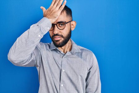 Foto de Middle east man with beard standing over blue background surprised with hand on head for mistake, remember error. forgot, bad memory concept. - Imagen libre de derechos