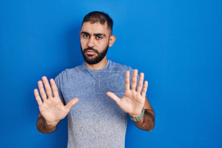Photo for Middle east man with beard standing over blue background moving away hands palms showing refusal and denial with afraid and disgusting expression. stop and forbidden. - Royalty Free Image