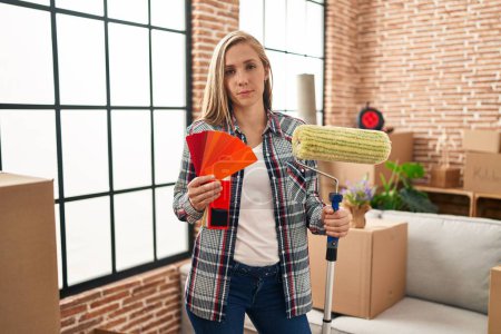 Photo for Young blonde woman holding roller painter and paint samples at new home relaxed with serious expression on face. simple and natural looking at the camera. - Royalty Free Image