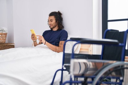 Téléchargez les photos : Young hispanic man disabled using smartphone sitting on bed at bedroom - en image libre de droit