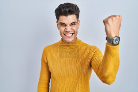 Photo for Young hispanic man standing over blue background angry and mad raising fist frustrated and furious while shouting with anger. rage and aggressive concept. - Royalty Free Image