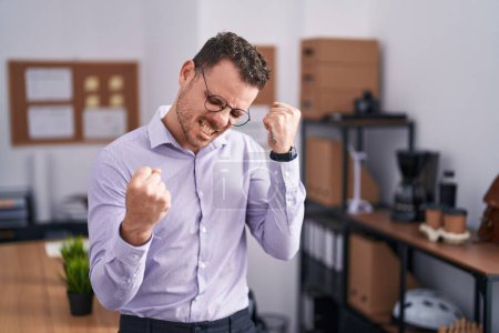 Photo for Young hispanic man at the office very happy and excited doing winner gesture with arms raised, smiling and screaming for success. celebration concept. - Royalty Free Image