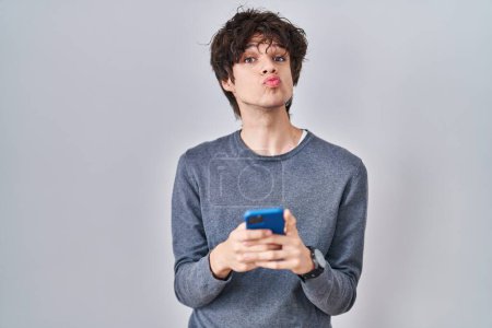 Photo for Young man using smartphone typing a message looking at the camera blowing a kiss being lovely and sexy. love expression. - Royalty Free Image