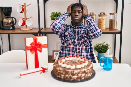 Foto de Hombre africano con rastas celebrando cumpleaños celebrando gran pastel de chocolate sufriendo de dolor de cabeza desesperado y estresado porque el dolor y la migraña. manos en la cabeza. - Imagen libre de derechos