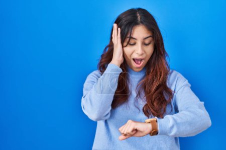 Téléchargez les photos : Jeune femme hispanique debout sur fond bleu regardant le temps de la montre inquiet, peur de se faire tard - en image libre de droit
