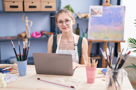 Téléchargez les photos : Jeune artiste femme caucasienne souriant confiant en utilisant un ordinateur portable au studio d'art - en image libre de droit