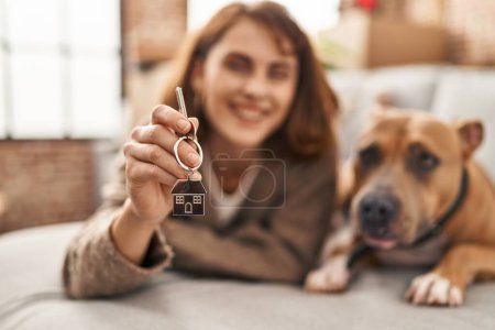 Téléchargez les photos : Jeune femme caucasienne tenant la clé de la nouvelle maison couchée sur le canapé avec chien à la nouvelle maison - en image libre de droit