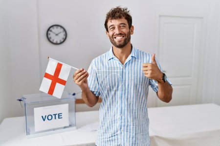 Foto de Joven hombre guapo en las elecciones de campaña política sosteniendo bandera de Inglaterra sonriendo feliz y positivo, pulgar hacia arriba haciendo excelente y señal de aprobación - Imagen libre de derechos