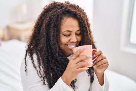 Foto de Mujer afroamericana bebiendo taza de café sentada en la cama en el dormitorio - Imagen libre de derechos