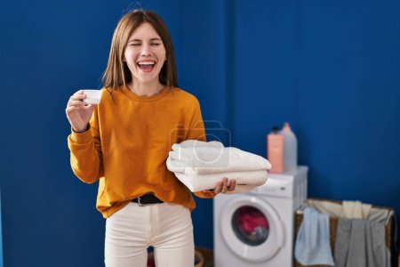 Foto de Joven morena sosteniendo detergente y ropa limpia sonriendo y riendo en voz alta porque divertida broma loca. - Imagen libre de derechos