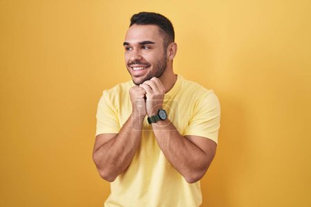 Téléchargez les photos : Jeune homme hispanique debout sur fond jaune riant nerveux et excité avec les mains sur le menton regardant vers le côté - en image libre de droit