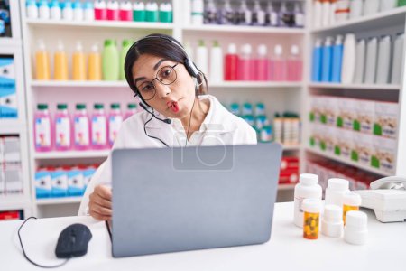 Foto de Young arab woman working at pharmacy drugstore using laptop making fish face with lips, crazy and comical gesture. funny expression. - Imagen libre de derechos