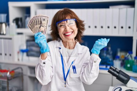 Foto de Mujer de mediana edad que trabaja en el laboratorio científico sosteniendo el dinero gritando orgulloso, celebrando la victoria y el éxito muy emocionado con los brazos levantados - Imagen libre de derechos