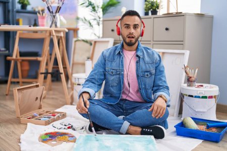 Photo for Young hispanic man painter sitting on the floor at art studio scared and amazed with open mouth for surprise, disbelief face - Royalty Free Image