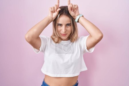 Téléchargez les photos : Jeune femme blonde debout sur fond rose faisant un geste drôle avec le doigt sur la tête comme des cornes de taureau - en image libre de droit