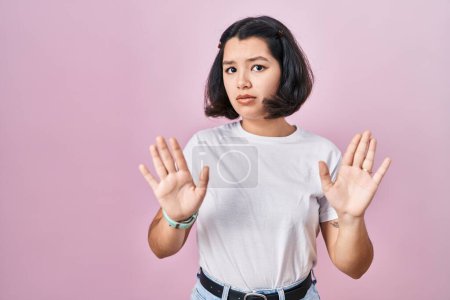 Photo for Young hispanic woman wearing casual white t shirt over pink background moving away hands palms showing refusal and denial with afraid and disgusting expression. stop and forbidden. - Royalty Free Image