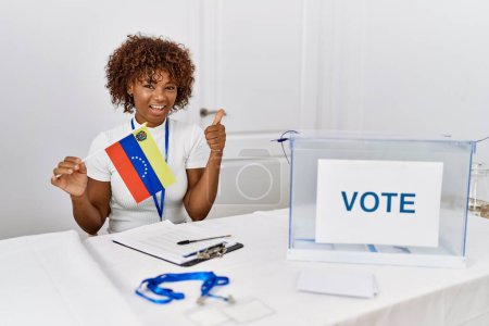 Foto de Mujer afroamericana joven en las elecciones de campaña política sosteniendo bandera de venezuela sonriendo feliz y positivo, pulgar hacia arriba haciendo excelente y signo de aprobación - Imagen libre de derechos