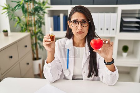 Photo for Young hispanic doctor woman holding heart and cbd oil depressed and worry for distress, crying angry and afraid. sad expression. - Royalty Free Image