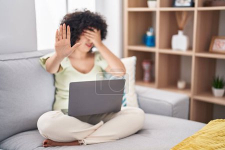 Photo for Young brunette woman with curly hair using laptop sitting on the sofa at home covering eyes with hands and doing stop gesture with sad and fear expression. embarrassed and negative concept. - Royalty Free Image