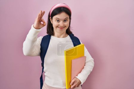 Foto de Mujer con síndrome de Down vistiendo mochila de estudiante y sosteniendo libros sonriendo positiva haciendo signo de ok con la mano y los dedos. expresión exitosa. - Imagen libre de derechos