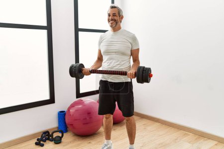 Foto de Hombre de pelo gris de mediana edad sonriendo confiado usando entrenamiento con pesas en el centro deportivo - Imagen libre de derechos