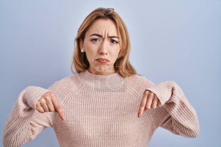 Photo for Hispanic woman standing over blue background pointing down looking sad and upset, indicating direction with fingers, unhappy and depressed. - Royalty Free Image