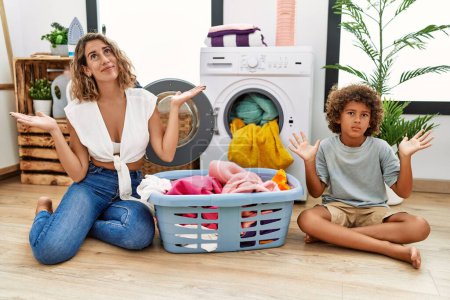 Photo for Young woman and son putting dirty laundry into washing machine clueless and confused expression with arms and hands raised. doubt concept. - Royalty Free Image