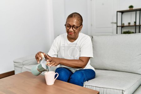 Foto de Mujer afroamericana mayor sonriendo confiada vertiendo café en la taza en casa - Imagen libre de derechos